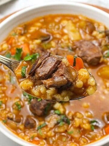 A close-up of a spoonful of beef barley soup held above a bowl. The soup contains chunks of beef, carrots, barley, and herbs in a thick broth. The bowl is on a wooden surface with sprigs of parsley nearby.