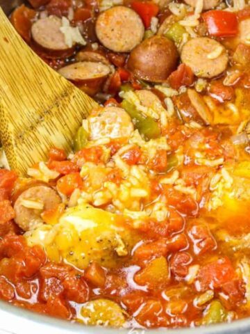 A pot filled with jambalaya, containing rice, sliced sausages, diced tomatoes, bell peppers, and spices, is being stirred with a wooden spoon—a perfect meal to save your life on busy nights.