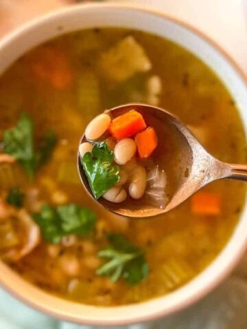 A hand holding a spoon filled with a vegetable soup containing carrots, white beans, and garnished with cilantro, positioned above a bowl of the same soup on a wooden surface.