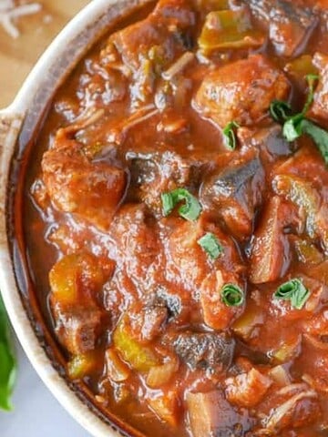 A ceramic dish filled with a hearty stew consisting of chunks of beef, tomatoes, bell peppers, and herbs. Fresh basil leaves are used as garnish. A slice of bread and grated cheese are visible in the background on a wooden board.