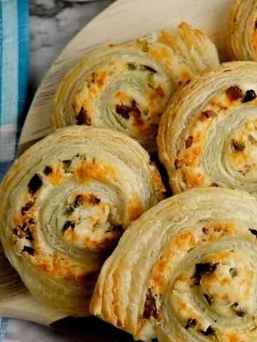 A wooden board holds several spiraled pastry rolls with a golden-brown crust, filled with cheese and speckled with herbs. These easy recipes are sure to be a hit. A blue checkered cloth is partially visible on the left side, inviting everyone to try one and beg you for the details.