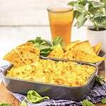 A baked spinach dip crowned with a golden parmesan crust rests in a dark rectangular baking dish. Beside it are crispy tortilla chips. In the background, a tall glass of iced drink and a potted green plant are visible, with fresh greens scattered around.