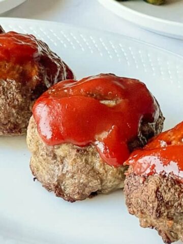 Four meatloaves are lined up on a white oval plate, each topped with a layer of red ketchup. The background shows a glimpse of another plate with assorted vegetables.