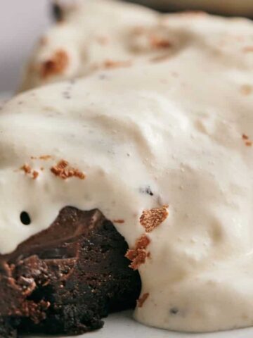 A slice of chocolate brownie with visible nuts, topped with a layer of creamy white icing, is placed on a white plate.