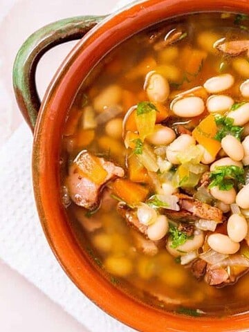 A clay bowl filled with bean soup, featuring white beans, chopped vegetables, and pieces of meat in a broth. The bowl is placed on a white textured cloth with a metal spoon to the side.