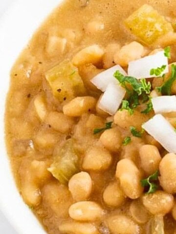 A close-up of a bowl of baked beans garnished with chopped onions and herbs. The beans are mixed with small pieces of green vegetables and sit in a thick, brown sauce.