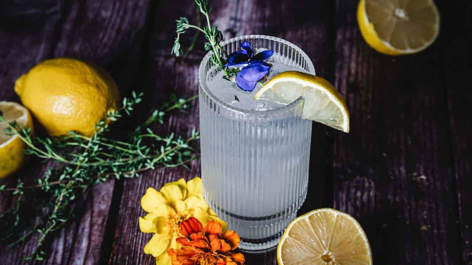 A tall, ribbed glass filled with a clear beverage, garnished with a lemon slice, a purple flower, and a sprig of herbs. Two whole lemons, one halved, some herbs, and two yellow flowers are scattered on the dark wooden surface.