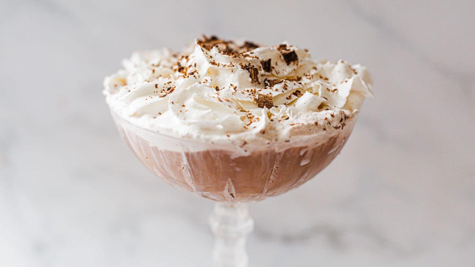 A chocolate dessert served in a glass bowl on a stand, topped with whipped cream and chocolate shavings, placed on a white marble surface.