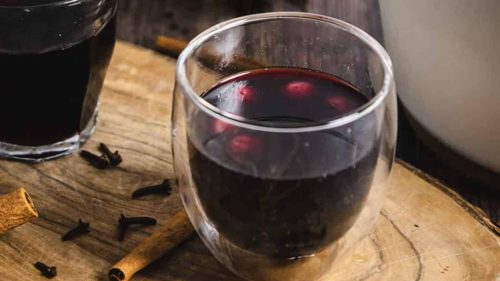 A glass of mulled wine sits on a wooden surface. The drink is garnished with a few floating berries. Nearby, there are whole cloves and a cinnamon stick on the wood. Another glass of dark liquid is partially visible in the background.