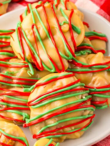 A plate of holiday-themed cookies is placed on a red and white checkered cloth. The cookies are drizzled with red and green icing over a caramel-colored base, surrounded by red and white candy beads.