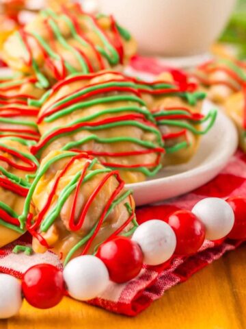 A plate of cookies drizzled with red and green icing is displayed on a red checkered cloth. Decorative red and white beads surround the platter. A glass of milk and some greenery are visible in the background.