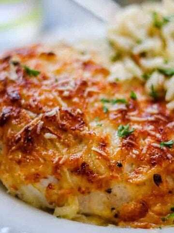 A plate of baked chicken breast with melted cheese and herbs, served with a portion of parsley-garnished rice. This cozy dinner idea features golden brown chicken and fluffy rice, all elegantly presented on a white plate—a perfect addition to your collection of oven recipes.