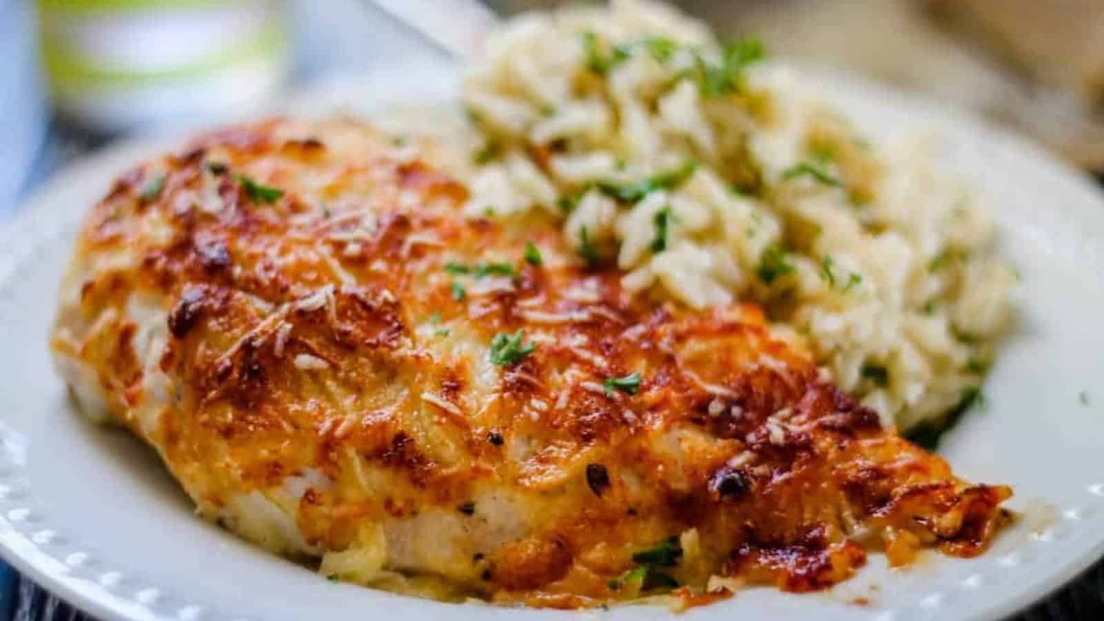 A plate of baked chicken breast with melted cheese and herbs, served with a portion of parsley-garnished rice. This cozy dinner idea features golden brown chicken and fluffy rice, all elegantly presented on a white plate—a perfect addition to your collection of oven recipes.