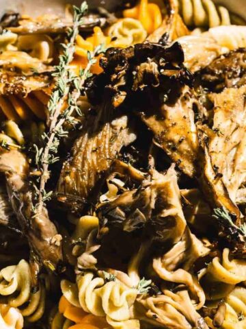 A bowl of tri-color fusilli pasta topped with sautéed mushrooms and garnished with sprigs of fresh thyme. The pasta and mushrooms are lightly seasoned, with the dish arranged on a wooden table surface.