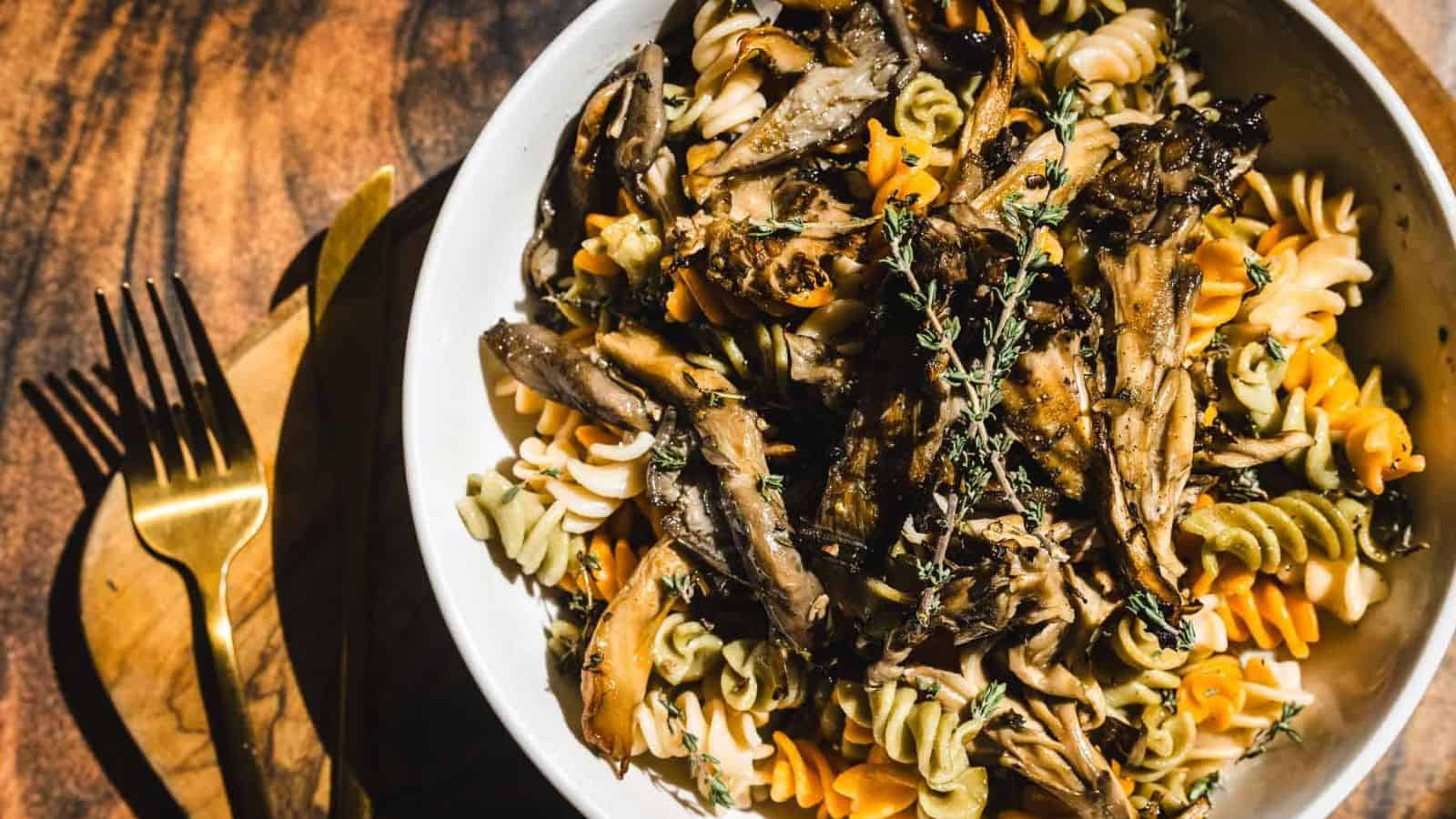 A bowl of multicolored fusilli pasta topped with cooked mushrooms and garnished with fresh thyme sprigs. The dish is served in a white bowl on a wooden surface.