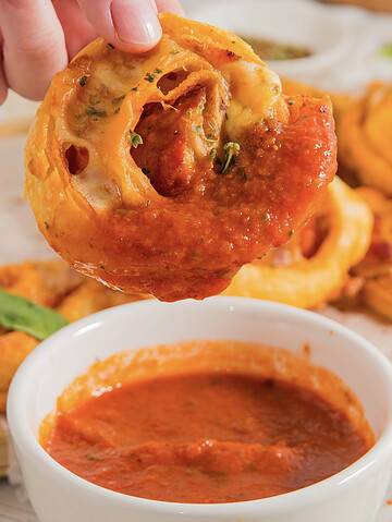 A hand holds a pepperoni pizza roll above a small white bowl filled with marinara sauce. The roll is partially dipped, showcasing its layers and toppings. More of these savory rolls are visible on a wooden board in the background.