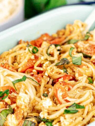 A dish of spaghetti mixed with diced tomatoes, herbs, and pieces of cheese, served in a rectangular light green dish. Reminiscent of Grandma's recipes, it holds a fork ready to twirl the pasta. A small bowl of grated cheese is visible in the background.