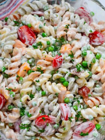 A glass bowl filled with creamy pasta salad, featuring tri-color rotini, cherry tomatoes, peas, and chopped herbs. A wooden spoon is partially visible next to the bowl on a marble surface. A striped cloth is nearby.