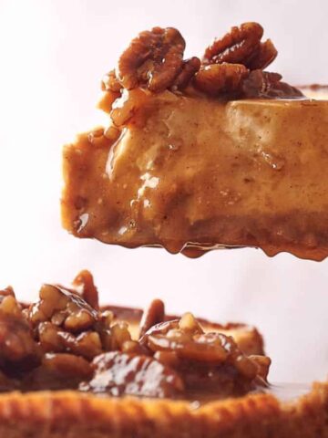 A slice of pecan-topped cheesecake is being lifted from a whole cheesecake. The dessert features a creamy filling and a crunchy pecan topping. The background is softly blurred.