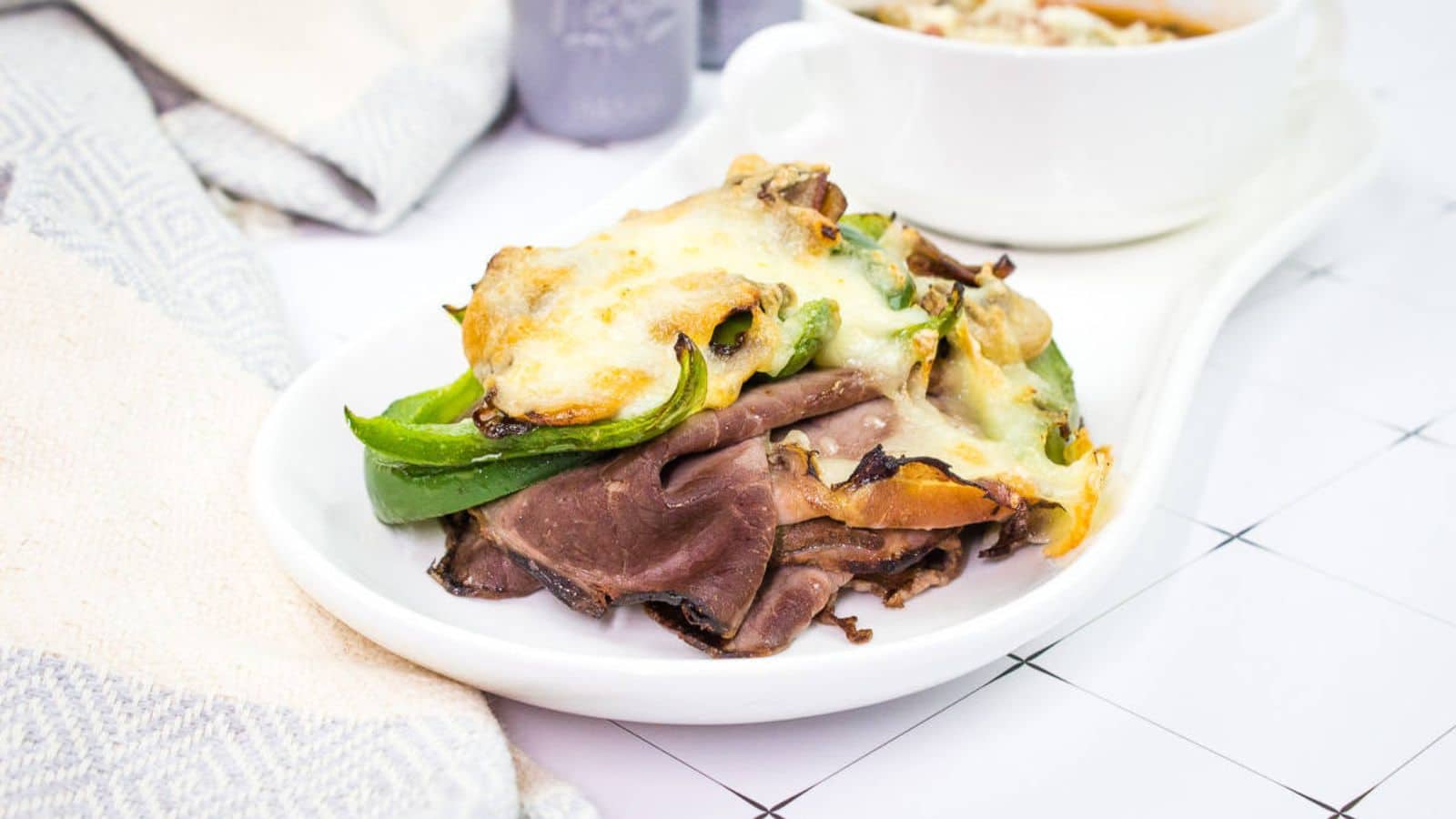 A white plate with a Philly Cheesesteak Stack and a bowl of soup.