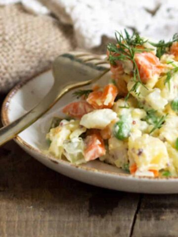 A plate of mixed vegetable salad featuring diced carrots, peas, and potatoes, garnished with fresh dill. A fork rests beside the plate on a rustic wooden table. A textured fabric is partially visible in the background.