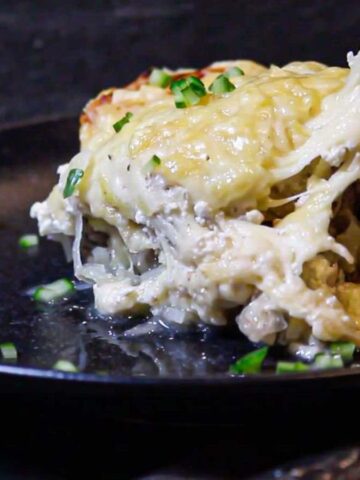 A serving of creamy, cheesy dish topped with chives on a dark plate offers the perfect solution for cozy dinners that are easy on your wallet. A portion is being lifted, showing melted cheese stretching above the dish. A fork is visible at the edge of the plate.