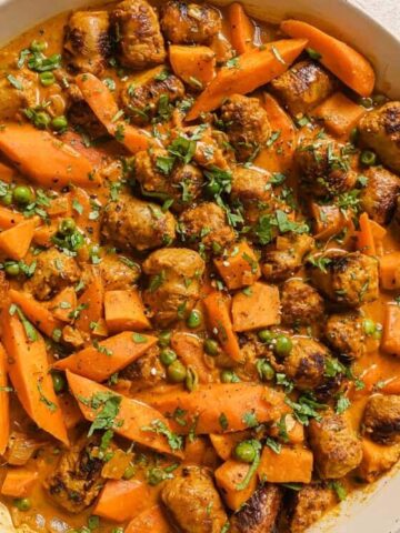 A ceramic dish filled with browned sausages, sliced carrots, and peas in a creamy orange sauce evokes the warmth of comfort food. Garnished with finely chopped herbs, it brings to mind the charm of forgotten recipes. A small bowl of herbs is visible at the bottom right corner.
