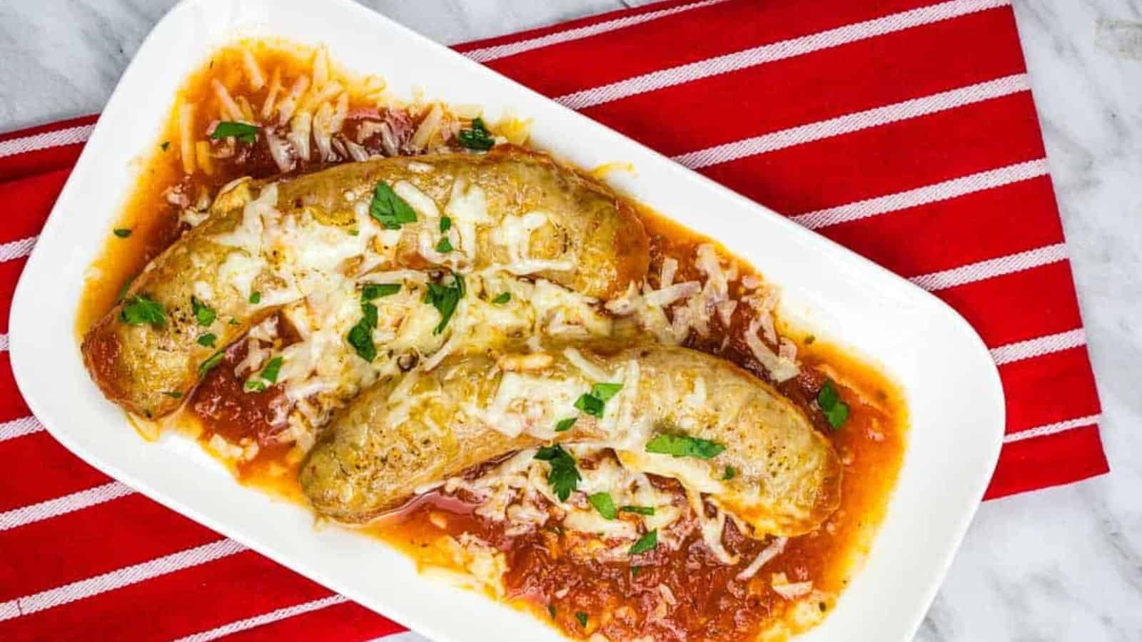 Two sausages topped with grated cheese and herbs sit on a bed of tomato sauce in a white rectangular dish. A red and white striped cloth is underneath.