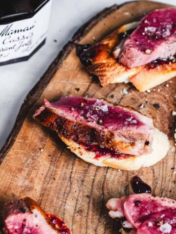 Sliced duck breast on toasted baguette slices with a berry sauce on a wooden board. A jar of fruit preserves is nearby.