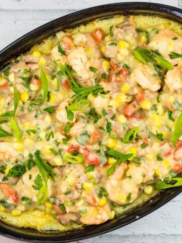 A black oval baking dish filled with a comforting shrimp and vegetable casserole sits on a whitewashed wooden surface. This delightful comfort food includes shrimp, corn, and tomatoes, all garnished with green herbs. A colorful cloth is nearby.