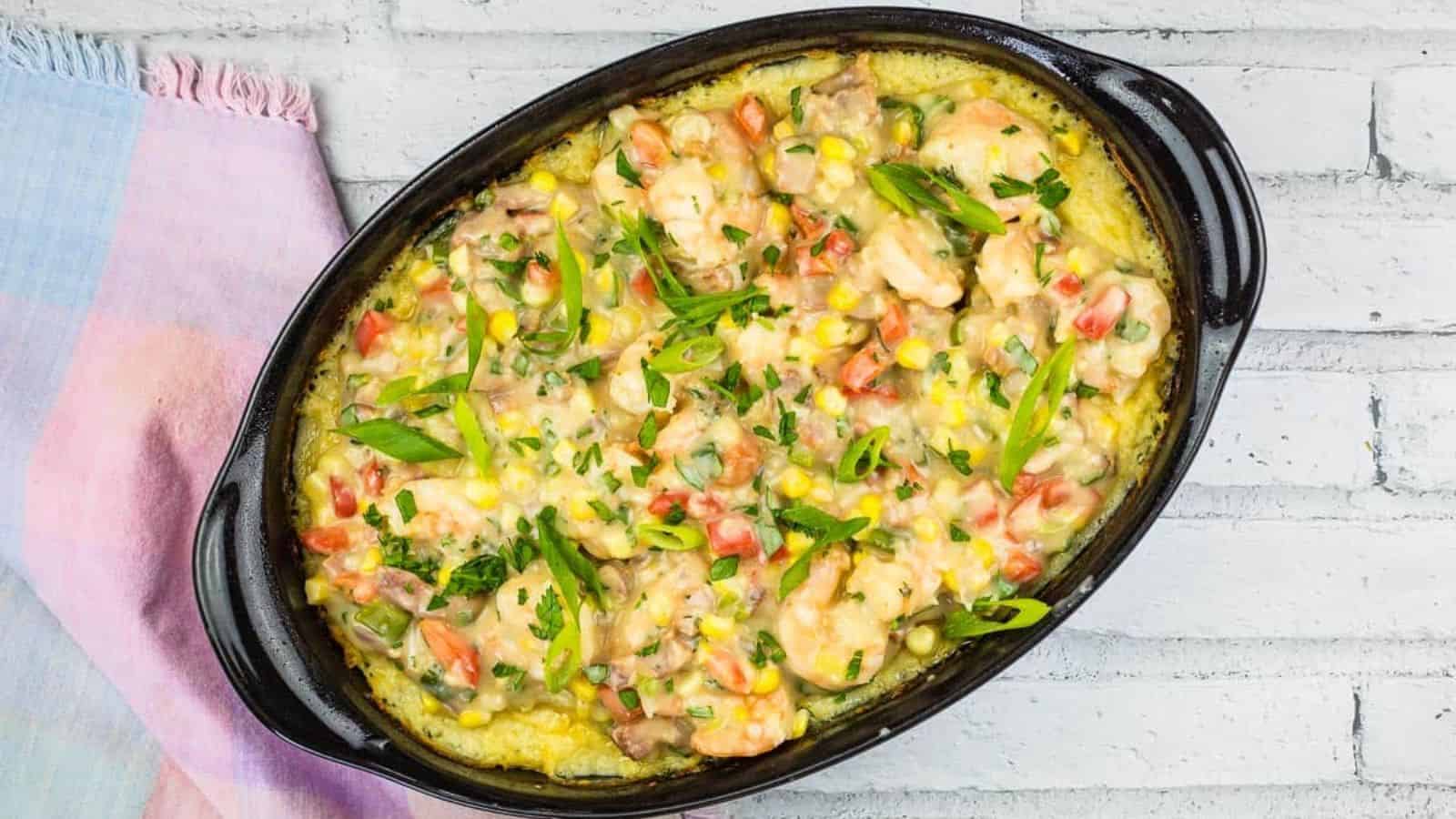 An oval baking dish contains a baked casserole topped with shrimp, sliced green onions, corn, diced tomatoes, and fresh parsley. A pastel-colored cloth napkin is placed beside the dish on a white wooden surface.