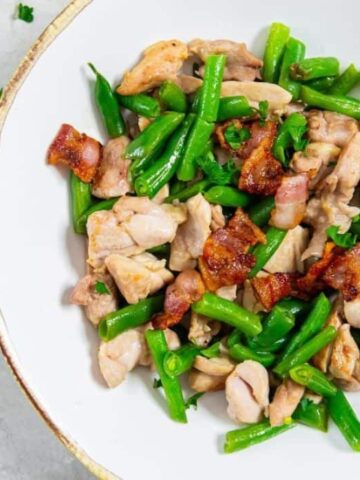 A dish with cooked chicken pieces, crispy bacon, and green beans is served in a white bowl. Silver forks are placed on the left side of the bowl. Fresh parsley leaves are scattered nearby on a light surface.