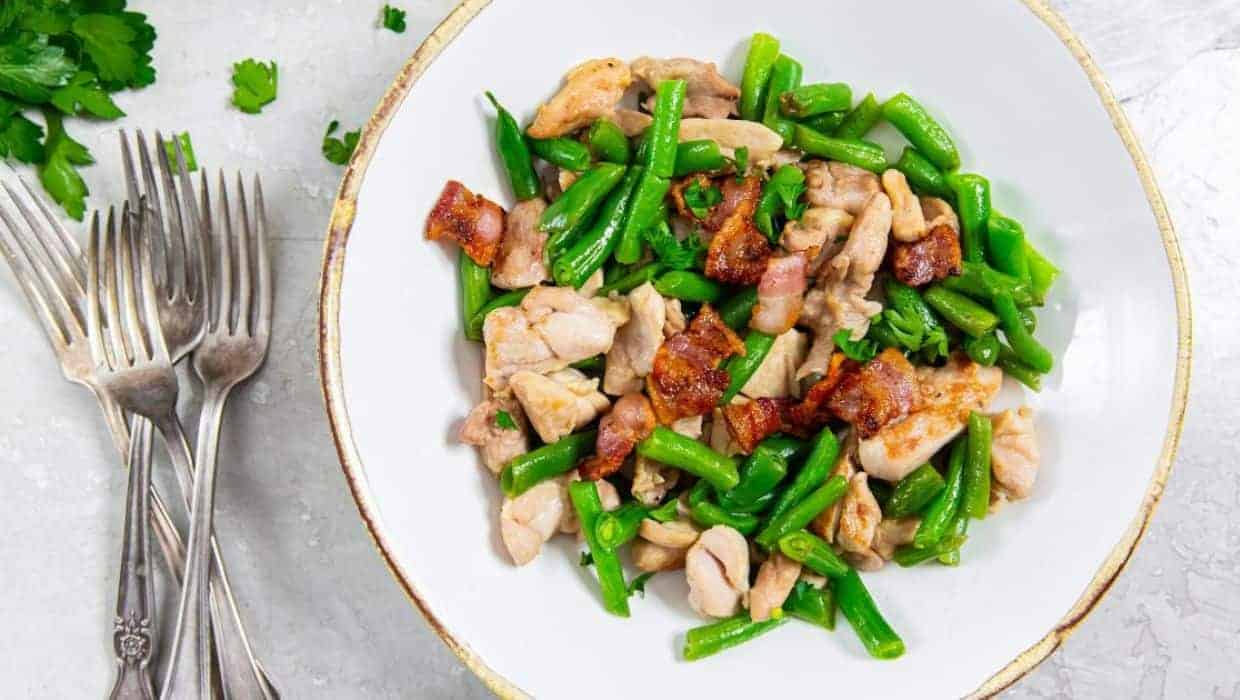 A dish with cooked chicken pieces, crispy bacon, and green beans is served in a white bowl. Silver forks are placed on the left side of the bowl. Fresh parsley leaves are scattered nearby on a light surface.