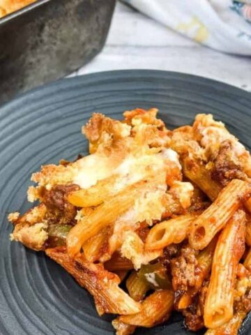 A serving of baked pasta casserole, featuring penne, ground meat, and melted cheese, is plated on a dark dish. In the background, a baking dish with more of this delicious recipe is visible alongside a floral napkin. A serving fork lies nearby.