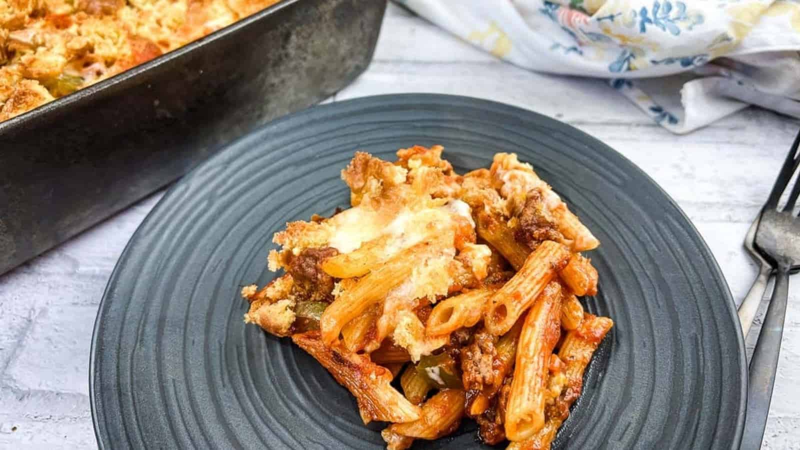 A serving of baked pasta casserole, featuring penne, ground meat, and melted cheese, is plated on a dark dish. In the background, a baking dish with more of this delicious recipe is visible alongside a floral napkin. A serving fork lies nearby.