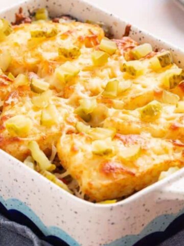 A white rectangular baking dish filled with a baked casserole topped with melted cheese, diced pickles, and golden-brown tater tots. The dish is placed on a folded blue cloth on a wooden surface. Plates are in the background.