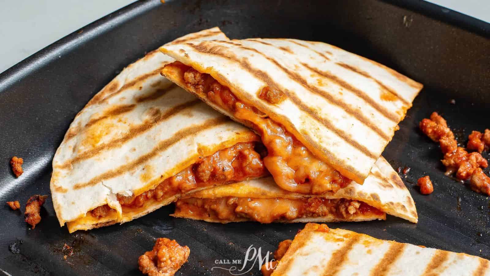 A grilled quesadilla filled with melted cheese and crumbled seasoned meat is cut into quarters and placed in a black skillet, embodying soul and comfort. Some meat pieces are scattered nearby, with the phrase "Call Me PMc" visible on the skillet's surface. A true hearty recipe to fill your belly.