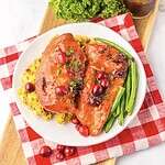 A white plate on a red checkered cloth holds two pieces of slow cooker cranberry chicken topped with cranberries and parsley, served with green beans and cornbread stuffing. A fresh parsley bunch sits in the background.