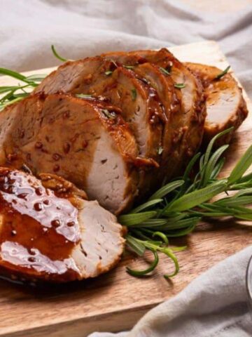 Sliced roasted pork tenderloin with a glaze is arranged on a wooden cutting board. Sprigs of rosemary are placed around the meat. A small bowl with additional glaze is nearby on a gray cloth.