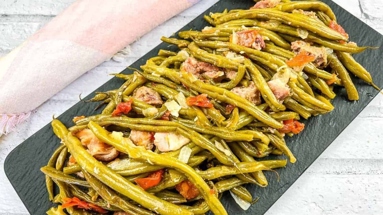 A slate platter showcases feel-good foods: cooked green beans mixed with chunks of meat and diced tomatoes. In the background, a folded cloth napkin rests on a pristine white surface, inviting a comforting dining experience.