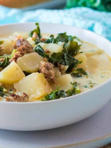 A white bowl of soul-soothing soup sits on a plate, with sausage, kale, and potato chunks in a creamy broth. This week's comfort food is elegantly set against a light blue cloth and leafy greens background.