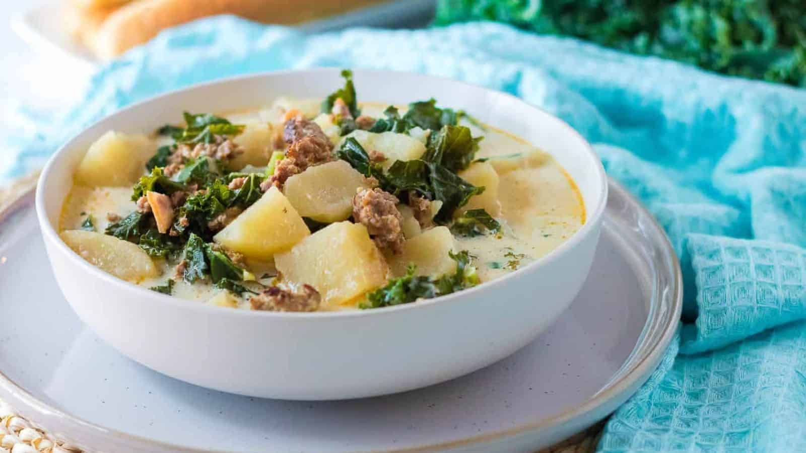 A white bowl of soul-soothing soup sits on a plate, with sausage, kale, and potato chunks in a creamy broth. This week's comfort food is elegantly set against a light blue cloth and leafy greens background.