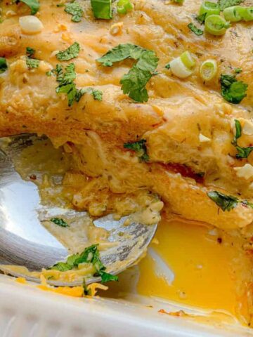 A close-up of a cheesy baked Mexican dish in a white rectangular baking dish. It has melted cheese, chopped green onions, and cilantro on top. A portion is missing, revealing layers inside, and a spoon rests in the dish.