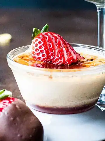 A small glass dish of crème brûlée topped with sliced strawberries sits on a wooden table. A chocolate-dipped strawberry is in the foreground. There are scattered heart-shaped candies and a partially visible wine glass nearby.