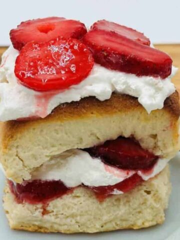 A slice of strawberry shortcake with layers of whipped cream and fresh strawberry slices sits on a white plate. A small jar filled with more strawberry slices is in the background. A fork is placed beside the cake.