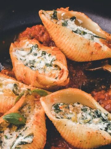 Stuffed pasta shells filled with spinach and ricotta are arranged on a black plate. The pasta rests on a layer of tomato sauce, and a gold fork is placed beside the shells. The dish is garnished with herbs.