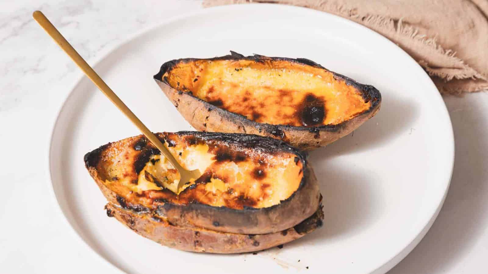 Two baked sweet potato halves on a white plate. The potatoes have a caramelized, slightly burnt surface, and one half is being scooped with a spoon.
