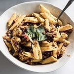 A delightful pasta dish featuring penne mixed with chicken, sun-dried tomatoes, and walnuts, garnished with fresh basil leaves. This creative take on a tofu pasta bake is served with a fork in the bowl against a light background.