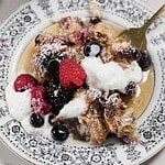 A plate with French toast bake, adorned with a variety of berries like raspberries, blackberries, and blueberries. Whipped cream and a sprinkle of powdered sugar grace the top. A gold fork completes the presentation on the patterned dish.