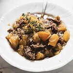 A white plate holds a savory Maple-Dijon Pot Roast stew with tender chunks of meat, potatoes, and vegetables in a rich brown broth. A sprig of fresh thyme garnishes the dish, adding a touch of elegance. A spoon rests in the bowl, inviting you to savor every bite.